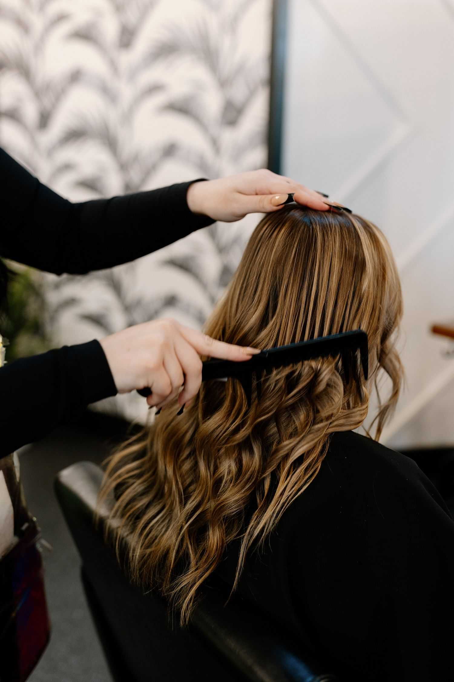 Stylist combing through wavy hair of a seated client in a salon.