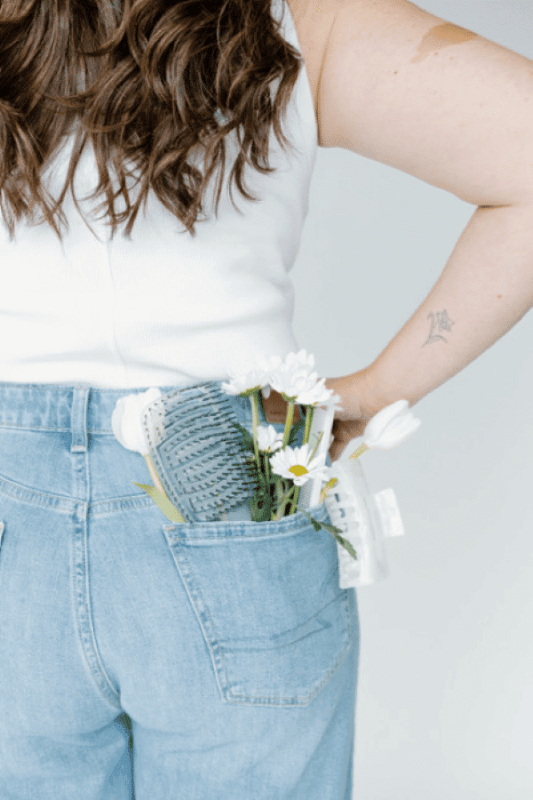 Woman with flowers and comb in back pocket of jeans, white top, arm tattoo visible.